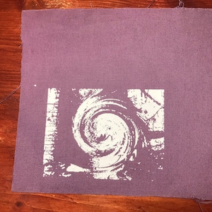 Puede incluir: Un parche de tela cuadrado y morado con un dise&ntilde;o blanco en espiral en el centro. El dise&ntilde;o se asemeja a una espiral o v&oacute;rtice. La tela tiene una apariencia texturizada y los bordes est&aacute;n sin terminar.