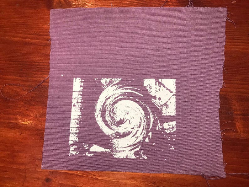 Puede incluir: Un parche de tela cuadrado y morado con un dise&ntilde;o blanco en espiral en el centro. El dise&ntilde;o se asemeja a una espiral o v&oacute;rtice. La tela tiene una apariencia texturizada y los bordes est&aacute;n sin terminar.