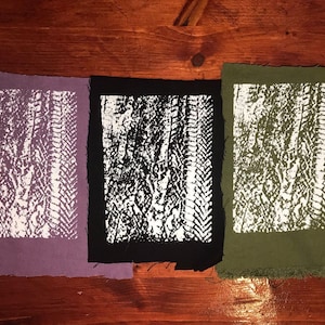 Può includere: Tre campioni di tessuto con un motivo bianco strutturato. I campioni sono di colore viola, nero e verde oliva. I bordi del tessuto sono sfilacciati e lo sfondo è una superficie in legno.