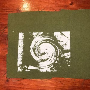 Puede incluir: Parche de tela verde oliva con un dise&ntilde;o de espiral blanco. El dise&ntilde;o es un remolino estilizado dentro de un marco rectangular. Los bordes del parche est&aacute;n sin terminar, con hilos deshilachados visibles. El parche est&aacute; sobre una superficie de madera.