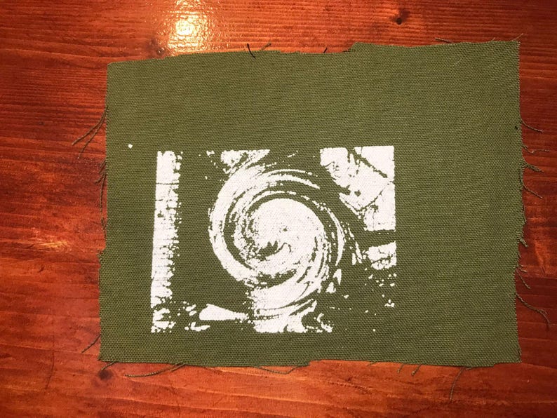 Puede incluir: Parche de tela verde oliva con un dise&ntilde;o de espiral blanco. El dise&ntilde;o es un remolino estilizado dentro de un marco rectangular. Los bordes del parche est&aacute;n sin terminar, con hilos deshilachados visibles. El parche est&aacute; sobre una superficie de madera.