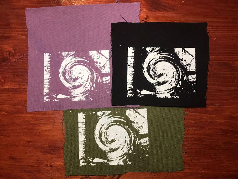 Puede incluir: Tres parches de tela en morado, negro y verde oliva, cada uno con un dise&ntilde;o de espiral blanco dentro de un borde rectangular blanco. Los parches est&aacute;n colocados sobre una superficie de madera.
