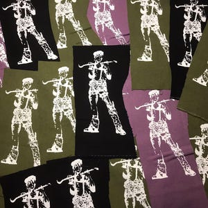 Könnte beinhalten: Eine Sammlung von Stoffaufnähern in verschiedenen Farben, darunter Schwarz, Olivgrün und Lila. Jeder Aufnäher zeigt eine weiße Grafik einer Person, die einen Bogen hält. Die Aufnäher sind rechteckig mit rohen, unfertigen Kanten.
