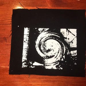Puede incluir: Parche cuadrado negro con un dise&ntilde;o blanco en espiral. El dise&ntilde;o est&aacute; contenido dentro de un rect&aacute;ngulo blanco. El parche tiene un borde sin rematar. El parche est&aacute; sobre una superficie de madera.