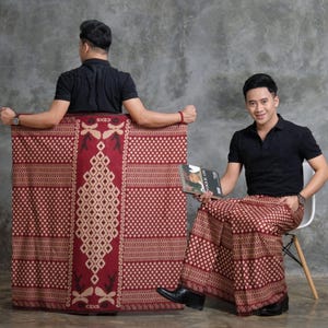 May include: A maroon and cream-colored sarong with a geometric pattern. The sarong is held up by a person wearing a black shirt. The sarong has the word "CENS" printed on it. The person is sitting on a white chair.