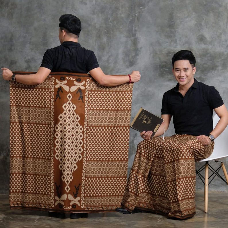 May include: Brown sarong with a geometric pattern and a central diamond design. The sarong is displayed by two people wearing black shirts. One person is holding a book. The sarong has the text "CENS".