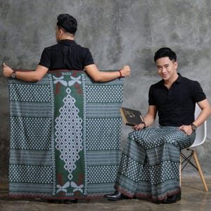 May include: Two people are shown wearing and holding a green and white patterned sarong. The sarong has a central diamond design and the word "CENS" is visible. One person is holding a book and sitting in a chair.
