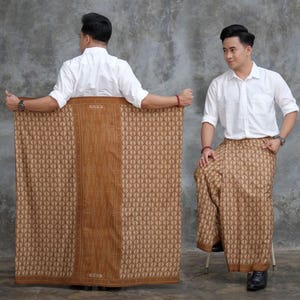 May include: A brown sarong with a geometric pattern and a central vertical panel. The sarong is held by a person wearing a white shirt. The word "CENS" is visible on the sarong.