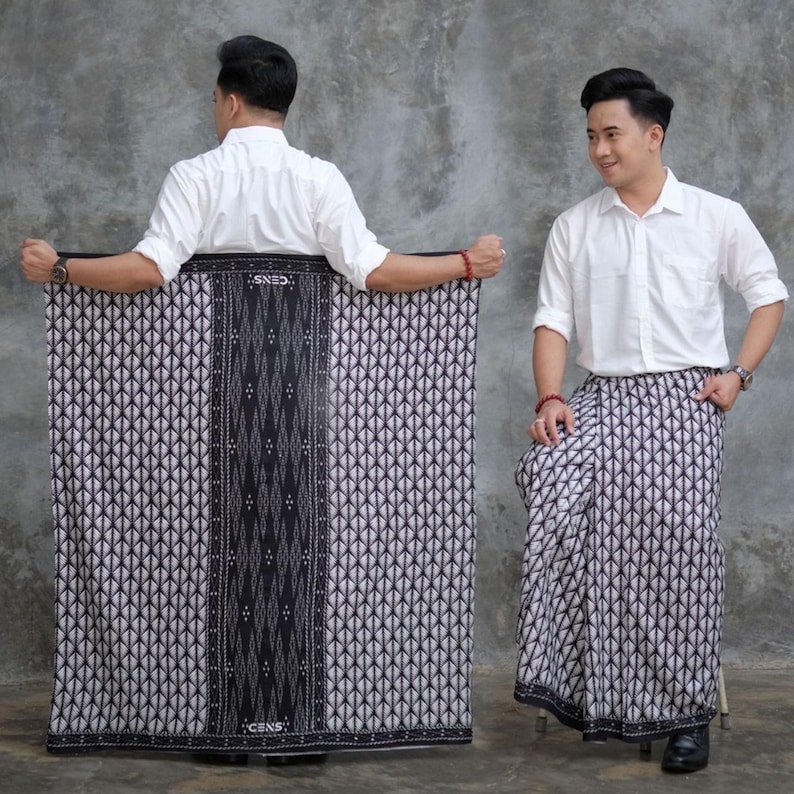 May include: A black and white patterned sarong, with a central dark vertical band. The sarong is worn by a person wearing a white button-down shirt. The sarong features a geometric design and the word "CENS" is visible.