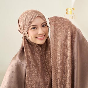 May include: A mocha-coloured hijab featuring a delicate floral lace overlay with a scalloped edge. The fabric has a subtle sheen and a repeating small flower pattern. The head covering is draped to frame the face, showcasing the intricate lace texture.