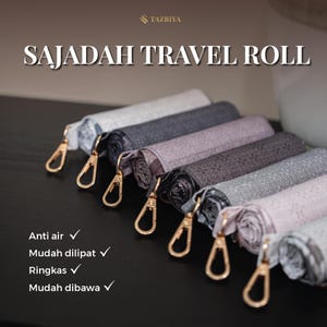 Op de afbeelding: Een collectie opgerolde gebedskleden met het label SAJADAH TRAVEL ROLL van TAZBIYA. Elk kleed heeft een uniek patroon in grijze, paarse en witte tinten en is voorzien van een goudkleurige metalen karabijnhaak aan een stoffen band.