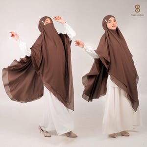 May include: Two women wearing brown hijabs and white dresses pose against a white backdrop. The hijabs drape over the shoulders and cover the head and face, with a small pin securing the fabric. The Sakinahijab logo is visible in the upper right corner.