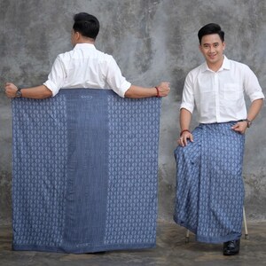 May include: A blue sarong with a geometric pattern, held by a person wearing a white shirt. The sarong has a darker blue central stripe and the text "SNED" at the top and bottom. The sarong is displayed in two different ways.