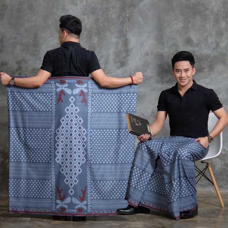 May include: Two people are shown wearing a blue sarong with a geometric pattern and red accents. The sarong has a diamond and dragonfly design. One person is holding a book. The sarong is a traditional garment.
