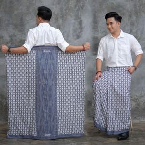 May include: A patterned sarong in shades of blue and white, held by a person wearing a white button-down shirt. The sarong features a geometric design with a central vertical stripe and the text "CENS". The fabric is draped to show the pattern.