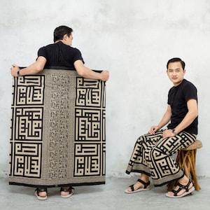 Könnte beinhalten: Zwei Männer sind abgebildet. Einer hält einen rechteckigen Stoff mit einem schwarz-beigen geometrischen Muster. Der andere Mann sitzt und trägt eine passende Hose. Beide Männer tragen schwarze T-Shirts und Sandalen. Der Stoff scheint ein Sarong zu sein.