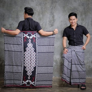 May include: A black and white patterned sarong with a diamond and geometric design. The sarong is held by a person wearing a black short-sleeved shirt. The sarong has a red border and the text "CENS" is visible.