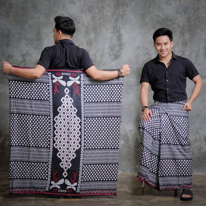 May include: A black and white patterned sarong with a diamond and geometric design. The sarong is held by a person wearing a black short-sleeved shirt. The sarong has a red border and the text "CENS" is visible.