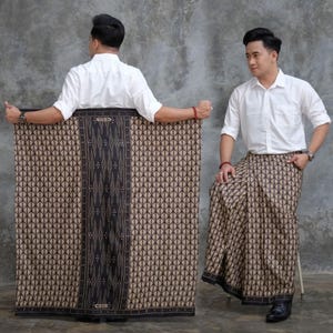 May include: A patterned sarong with a geometric design in shades of brown and black. The sarong is held by a person wearing a white shirt. The sarong has a dark border with the text "GENS".