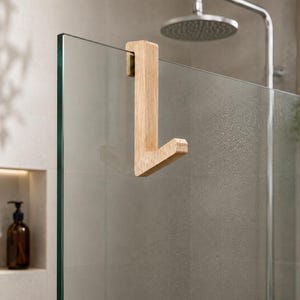 Könnte beinhalten: Ein hellfarbiger Holzhaken mit L-förmigem Design, der mit einem flachen oberen Teil über den Rand einer Glasduschwand gehängt wird. Der Haken hat eine glatte Oberfläche mit natürlicher Holzmaserung und eine stabile, kantige Konstruktion.