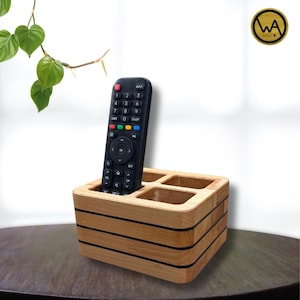Könnte beinhalten: Ein Fernbedienungshalter aus Holz mit vier Fächern. Der Halter besteht aus hellem Holz mit dunklen horizontalen Streifen. Eine schwarze Fernbedienung befindet sich in einem der Fächer. Der Halter hat ein modernes Design.