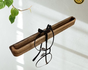 May include: A wooden eyeglasses holder mounted on a white wall. The holder is a long, rectangular trough with rounded edges, holding a pair of black-framed glasses. A small, round wooden logo is visible in the upper right corner. Green leaves are in the upper left corner.
