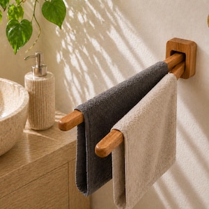 Könnte beinhalten: Handtuchhalter aus Holz mit zwei Handtüchern, eines grau und eines beige. Der Halter ist an einer weißen Wand befestigt. Ein Seifenspender und ein Waschbecken sind im Hintergrund zu sehen. Das Logo "WA" befindet sich an der Wand.
