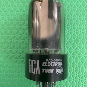 May include: A clear glass vacuum tube with a black cylindrical base. The base features white printed text that reads RCA RADIOTRON ELECTRON TUBE and includes the RCA logo. The internal metal components are visible through the glass dome.