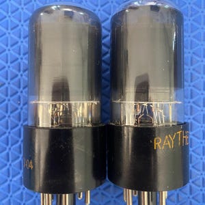 May include: Two vintage vacuum tubes with black bases and clear glass enclosures. The bases have multiple pins for connection. One tube is marked "7-04", and the other is marked "RAYTHEON". The tubes are cylindrical and appear to be for electronic equipment.