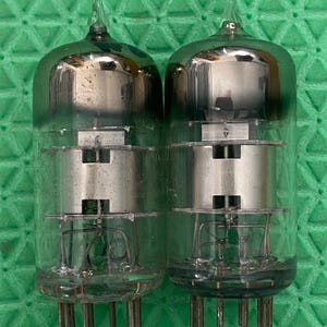 May include: Two vintage vacuum tubes with clear glass enclosures and silver internal components. Each tube has a pointed top and multiple pins at the base. The tubes are designed for electronic devices.