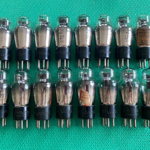 Puede incluir: Una colección de válvulas de vacío antiguas, cada una con una bombilla de vidrio y una base negra con pines. Los tubos están etiquetados como "27" y "SYLVANIA". Algunos tubos tienen etiquetas de papel. El fondo es verde liso.