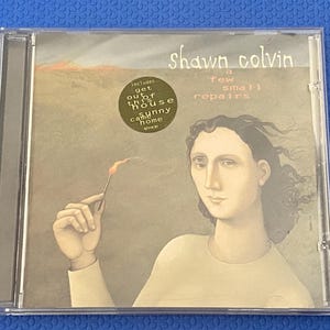 Puede incluir: Una caja de CD con el álbum "a few small repairs" de Shawn Colvin. La portada muestra a una persona sosteniendo un fósforo encendido. Incluye una pegatina que dice "Includes get out of this house sunny came home".