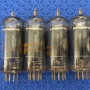 May include: Four vintage vacuum tubes with glass enclosures and multiple pins at the base. The tubes have a cylindrical shape with a rounded top. Some tubes have visible text. The background is a blue, textured surface.