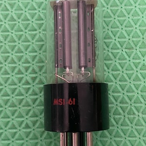 May include: A classic vacuum tube, featuring a transparent glass bulb and a black base. The base is marked with "MSI-61" in red lettering. The tube's internal structure is visible. The background is a green surface.