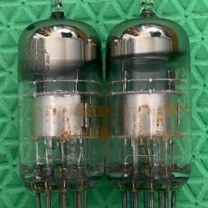 May include: Two vintage vacuum tubes with clear glass bulbs and silver-colored internal components. Each tube has a rounded top with a small glass protrusion and a series of pins at the base. The tubes are set against a green background.