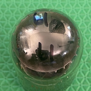 May include: A clear glass vacuum tube with a rounded top featuring a metallic silver getter coating on the interior. The number 61 is written in black ink on the exterior of the glass dome, which reflects the surrounding environment.