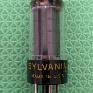 May include: A vintage Sylvania vacuum tube with a clear glass enclosure and a black base. The base has the text "SYLVANIA" and "MADE IN U.S.A." in gold lettering. The tube has multiple pins at the bottom.