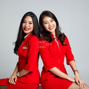 May include: Two women wearing matching red uniforms with short sleeves and skirts. They are smiling and sitting back-to-back. The uniforms have a zipper down the front and small badges. The background is a plain gray.
