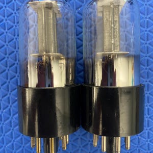 May include: Two vacuum tubes featuring clear glass envelopes and black cylindrical bases. Each tube contains internal metal components, including a central vertical structure and horizontal plates, with multiple metal pins extending from the bottom.