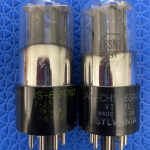 May include: Two vintage vacuum tubes with clear glass bulbs and black bases. The tubes have silver internal components and are labelled "6SN7GT" and "SYLVANIA". The bases have multiple pins for connection. The tubes are on a blue surface.