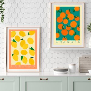 May include: Two framed prints with a white background and a light orange border. The print on the left features a yellow background with a pattern of lemons. The print on the right features a teal background with a pattern of oranges. The text "LEMONS" is at the bottom of the left print and the text "ORANGES" is at the bottom of the right print.