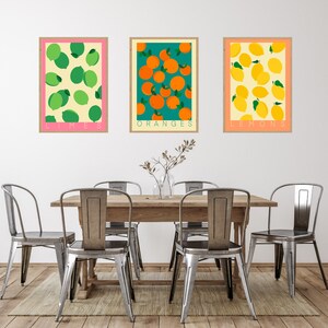 May include: Three framed prints of fruit illustrations. The top print is pink with green limes and the text "LIMES". The middle print is green with orange oranges and the text "ORANGES". The bottom print is orange with yellow lemons and the text "LEMONS".