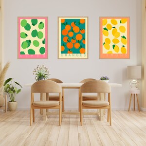May include: Three framed prints of fruit illustrations. The prints are hung on a white wall above a wooden table with two chairs. The prints feature limes, oranges, and lemons.