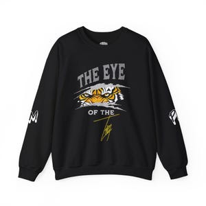 Puede incluir: Sudadera negra con el texto "THE EYE OF THE TIG" en una fuente desgastada. El diseño presenta el ojo de un tigre asomando a través de un efecto de papel rasgado. Las mangas tienen las letras "M" y "P".