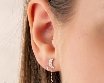 May include: A silver earring featuring a crescent moon design with embedded crystals. A delicate chain suspends a small star-shaped charm below the moon. The earring is worn on the ear.