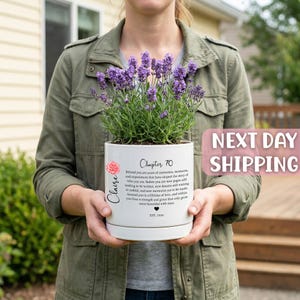 May include: A white ceramic planter containing a lavender plant with purple flowers. The planter features the text 'Chapter 70', the name 'Claire', a small pink flower graphic, a heart icon, and the text 'EST. 1956' printed on the front surface.