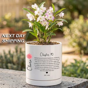 May include: A white ceramic planter containing a blooming orchid with light purple and white petals. The planter features black text reading Chapter 70, the name Claire, a pink flower graphic, a heart icon, and the text EST. 1956 with a long quote.