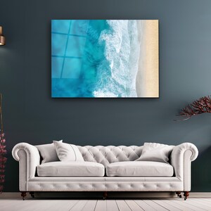 May include: A white couch with two pillows sits in front of a blue wall with a framed print of an aerial view of ocean waves crashing on a sandy beach.
