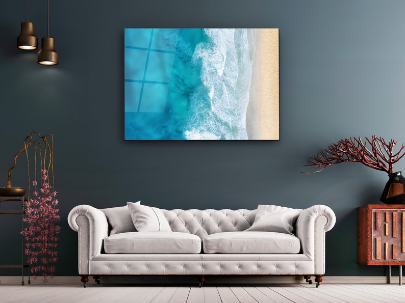 May include: A white couch with two pillows sits in front of a blue wall with a framed print of an aerial view of ocean waves crashing on a sandy beach.