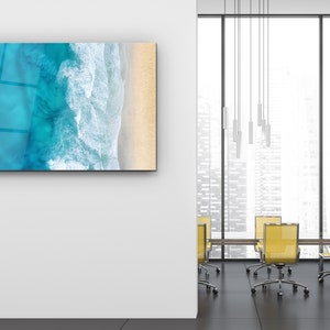 May include: A modern office space with a large window overlooking a city skyline. The room features a glass wall with a framed photograph of an aerial view of ocean waves crashing on a sandy beach. The room is furnished with a wooden table and yellow chairs.